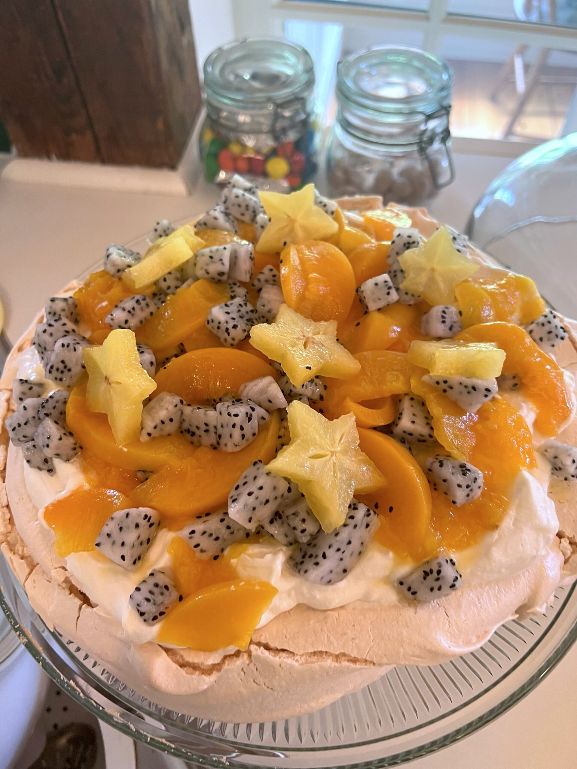 pavlova dessert with peaches, star fruit, dragon fruit, and whipped cream on a glass cake stand